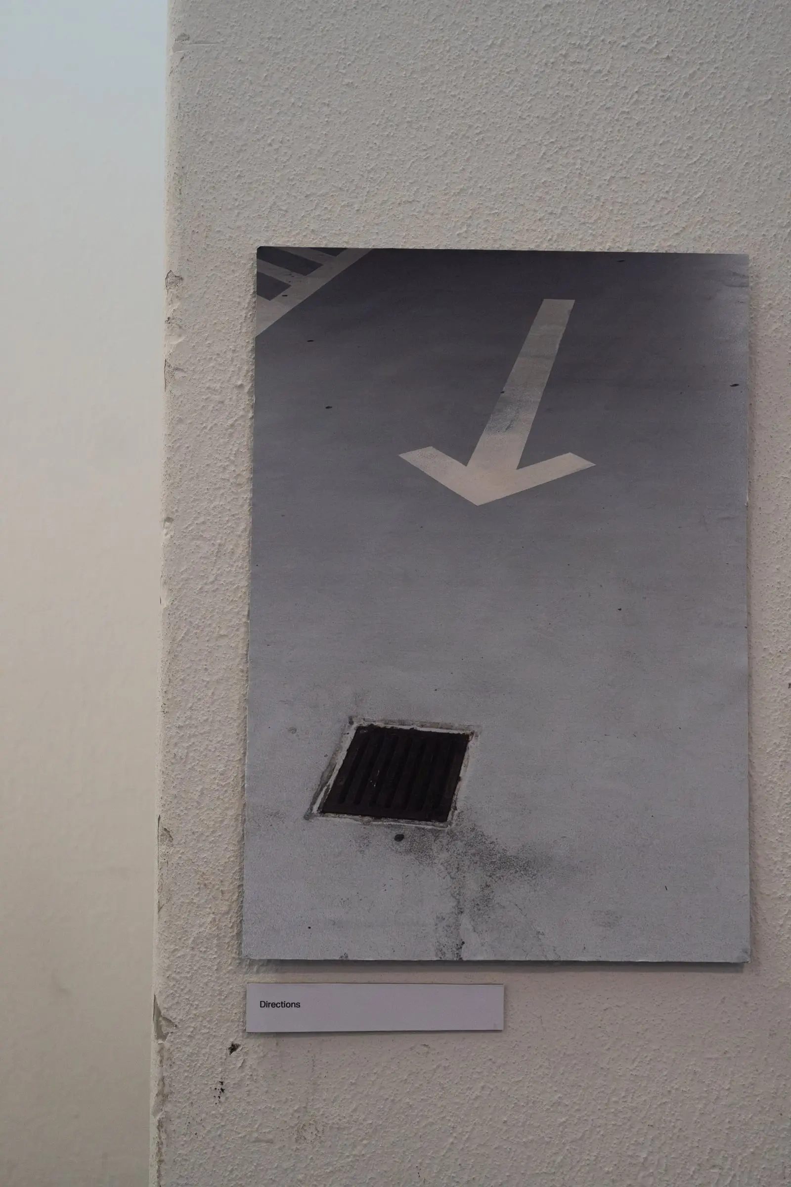 A print of a road arrow pointing at a drain, hung on a white wall