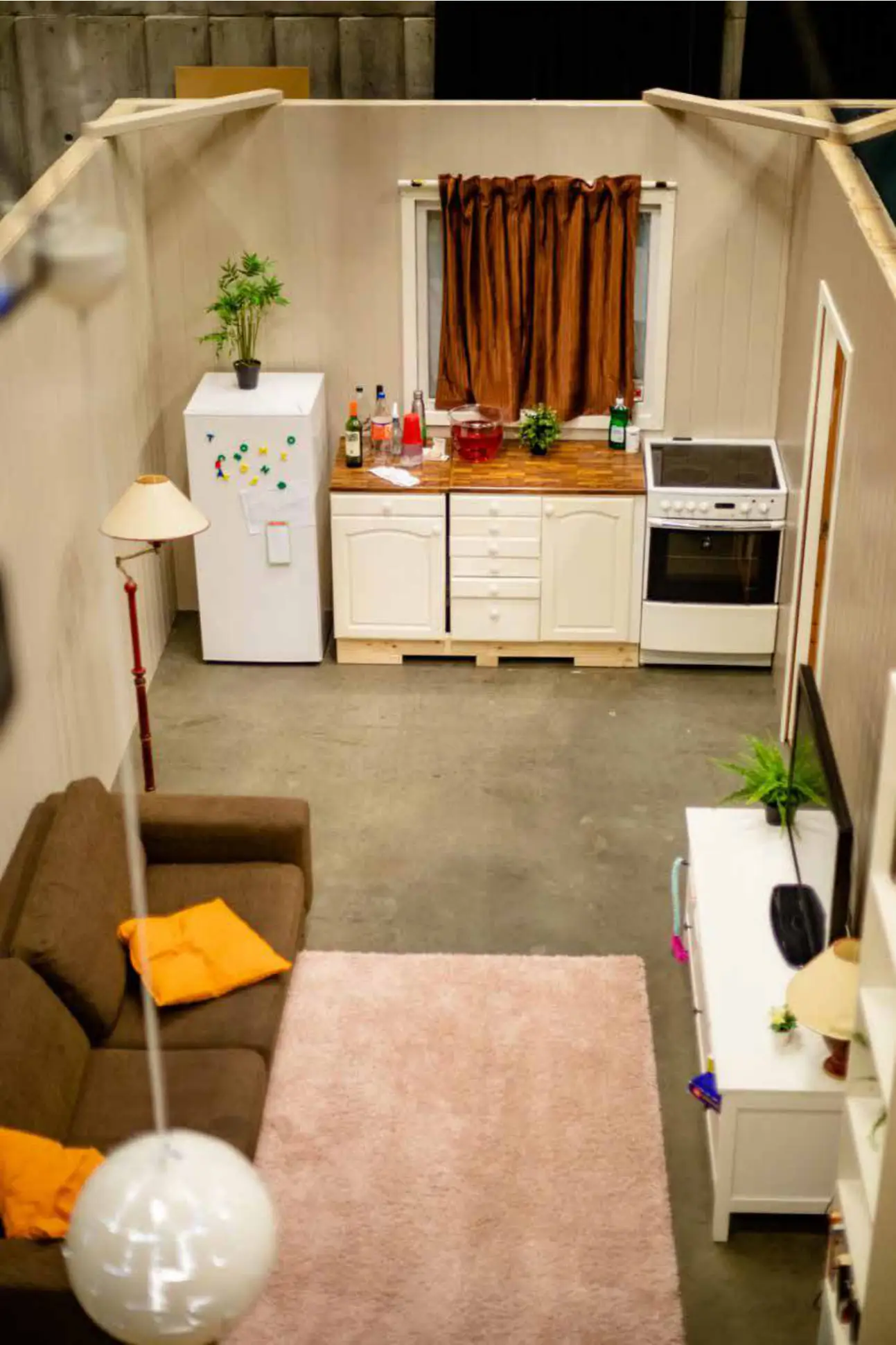 Furnished apartment set built inside a theatre, seen from above: kitchen, sofa, rug and lamps
