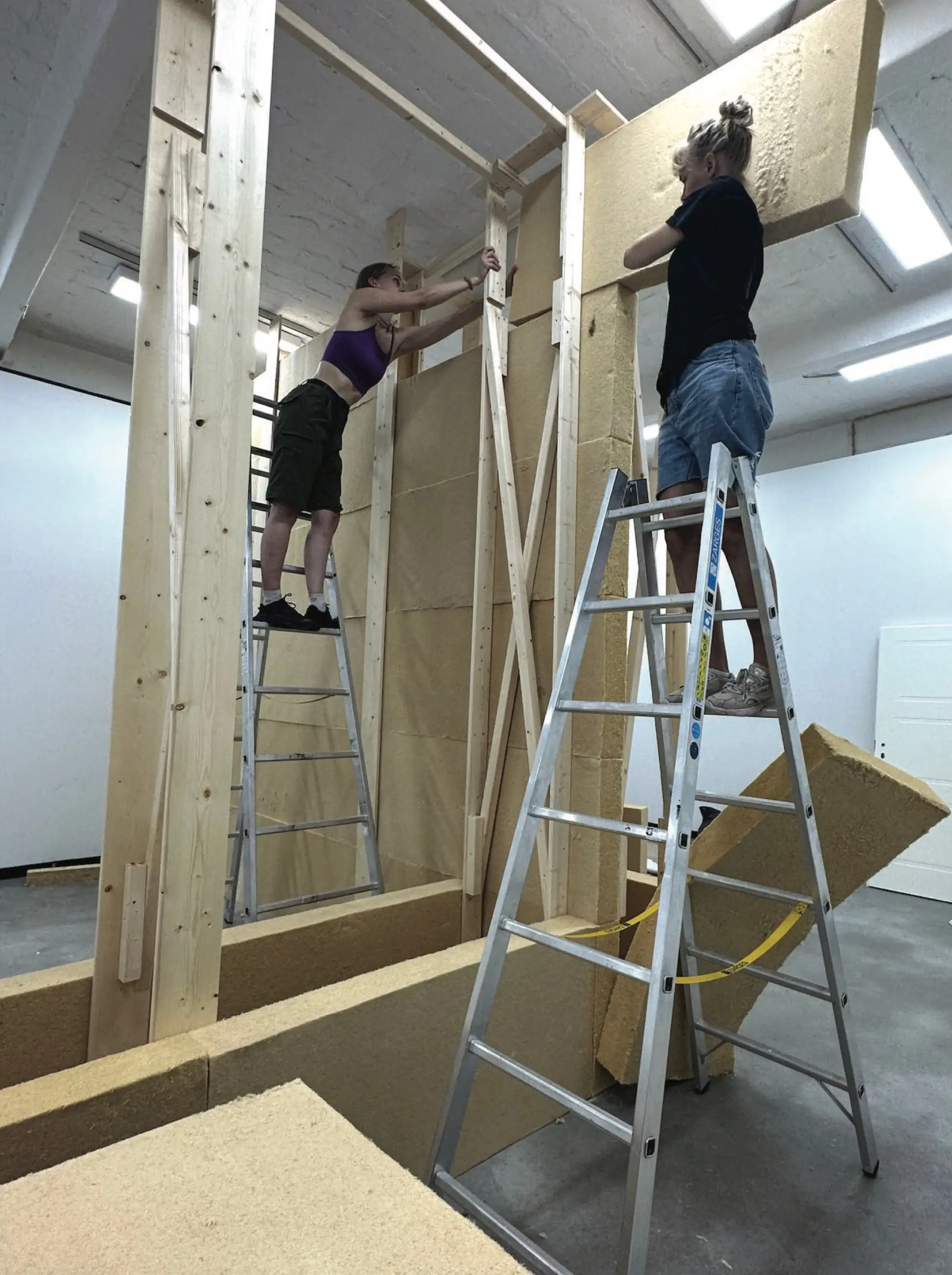 Two people on stepladders fixing soft wood-fibre insulation boards to a timber stud frame in a white gallery
