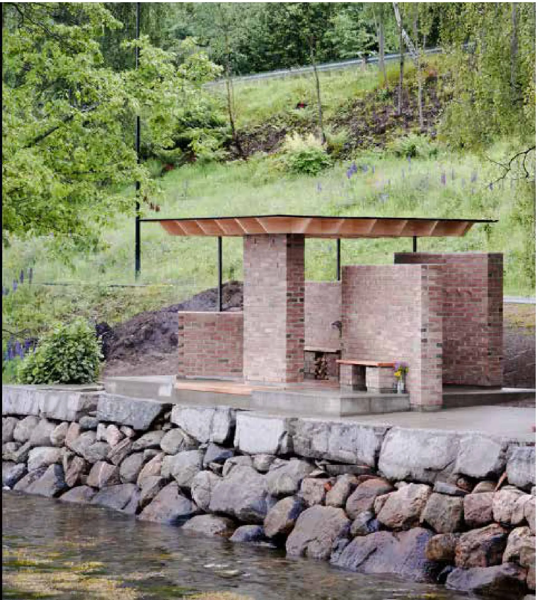 Brick and timber pavilion standing on a stone wall above a river, with a green slope behind