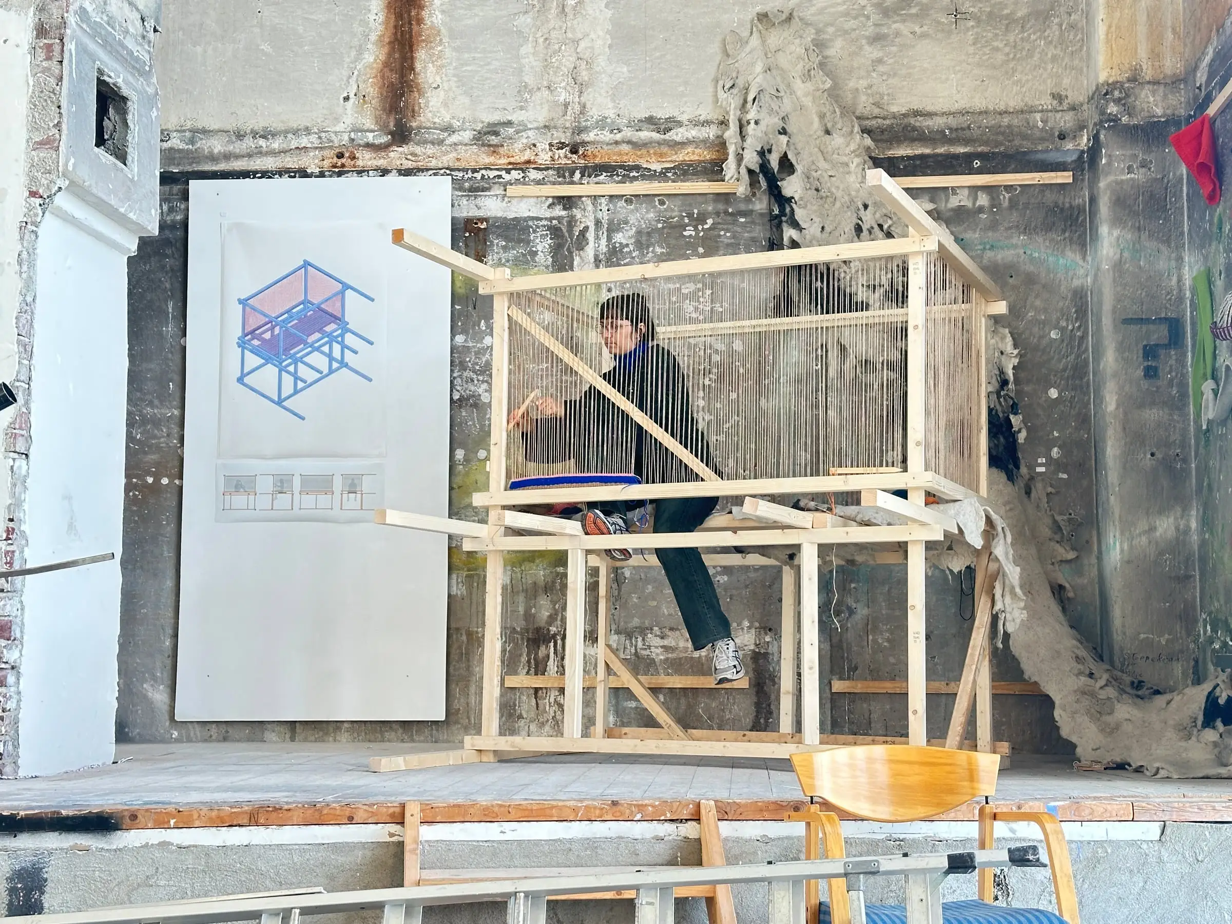 A timber frame built in a raw room, someone sitting inside it at a bench, a drawing of the loom pinned to a board beside it