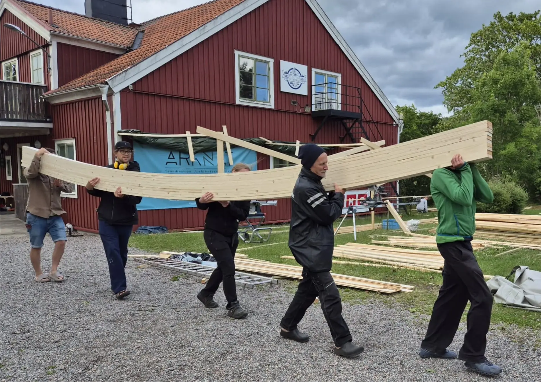 Five people carrying long curved timber beams across a gravel yard in front of a red house