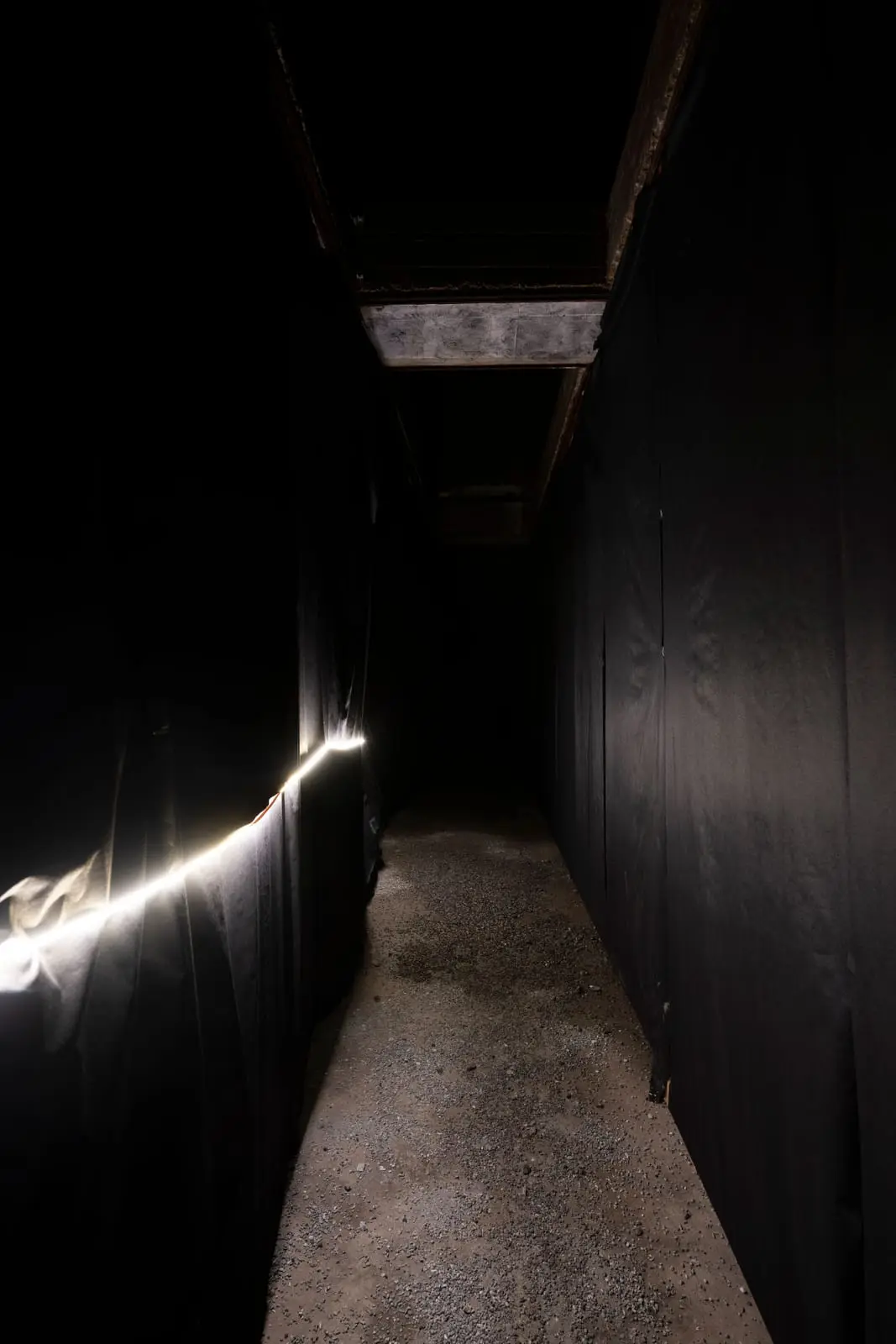 Narrow corridor between walls of black fabric, a strip of light running along one side over a gravel floor