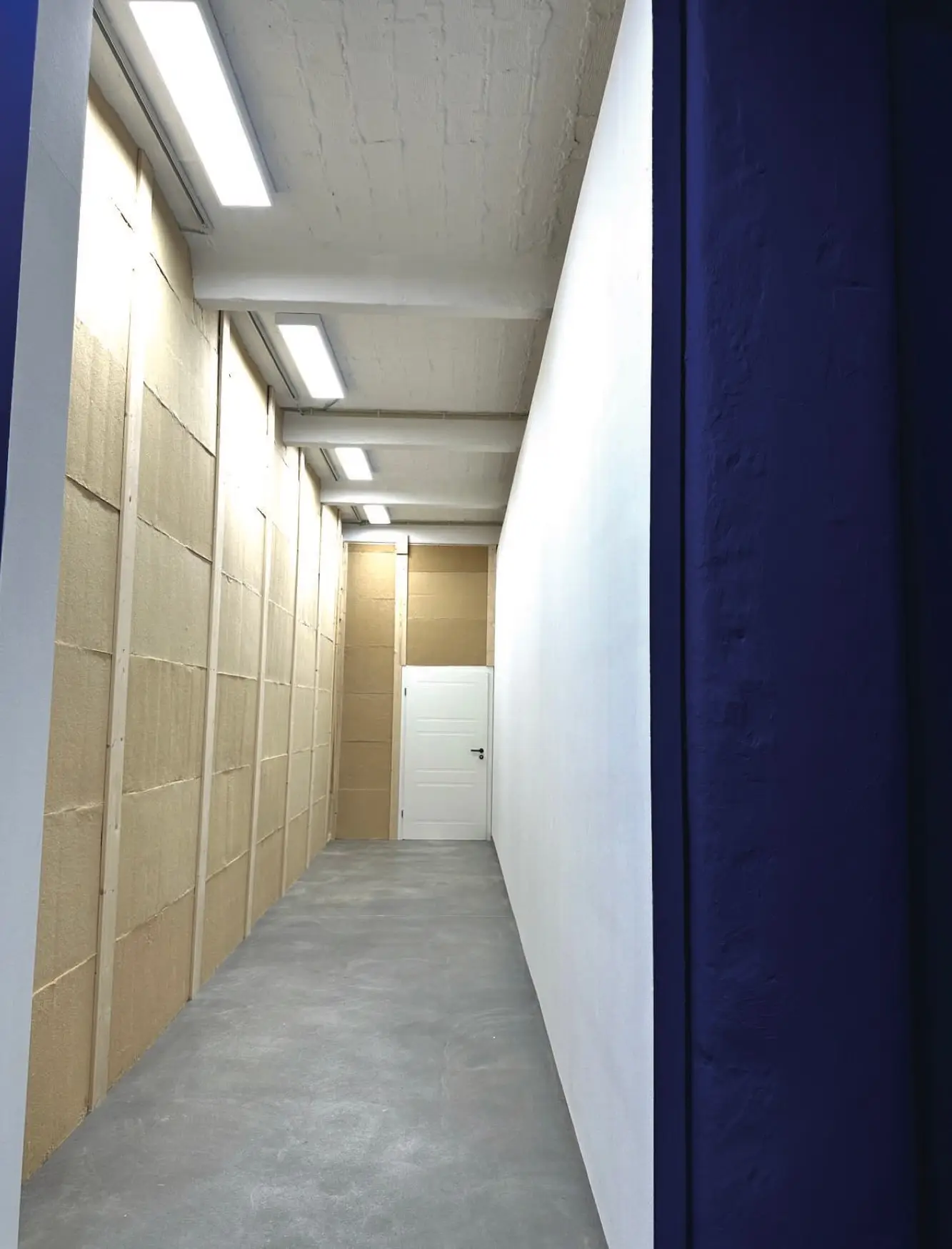 A long corridor lined with wood-fibre insulation boards between timber studs, a white door at the far end