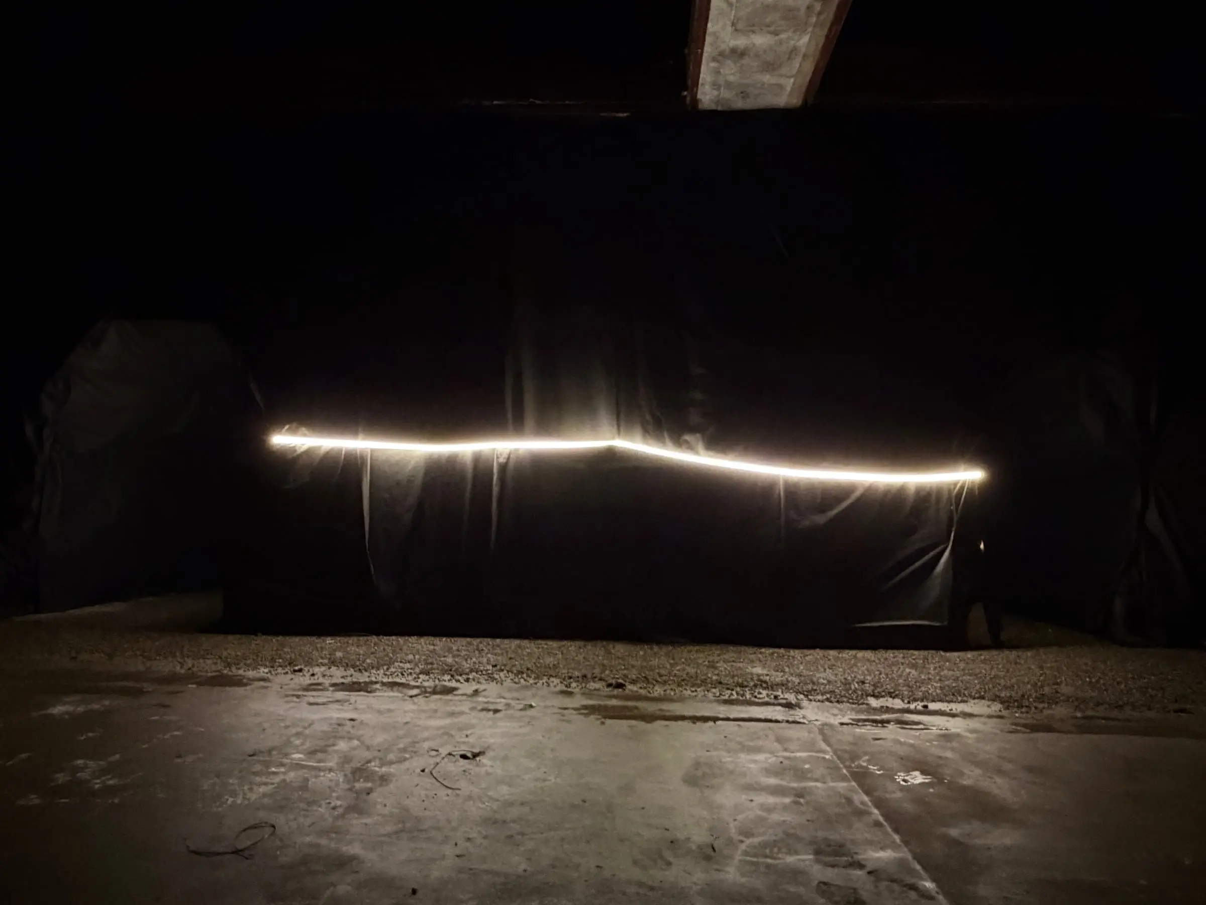 A long wall of black fabric with a strip of light along it, seen across an empty concrete floor