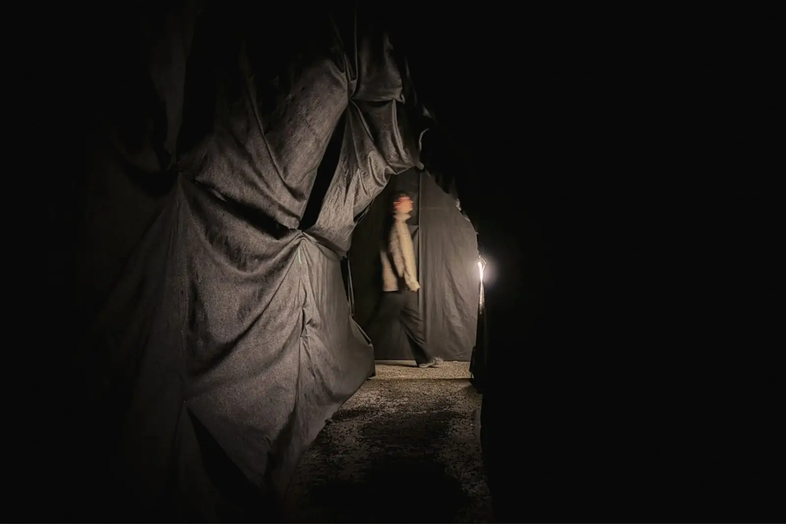 A visitor stepping through a torn opening in heavy black cloth, lit from one side, gravel underfoot