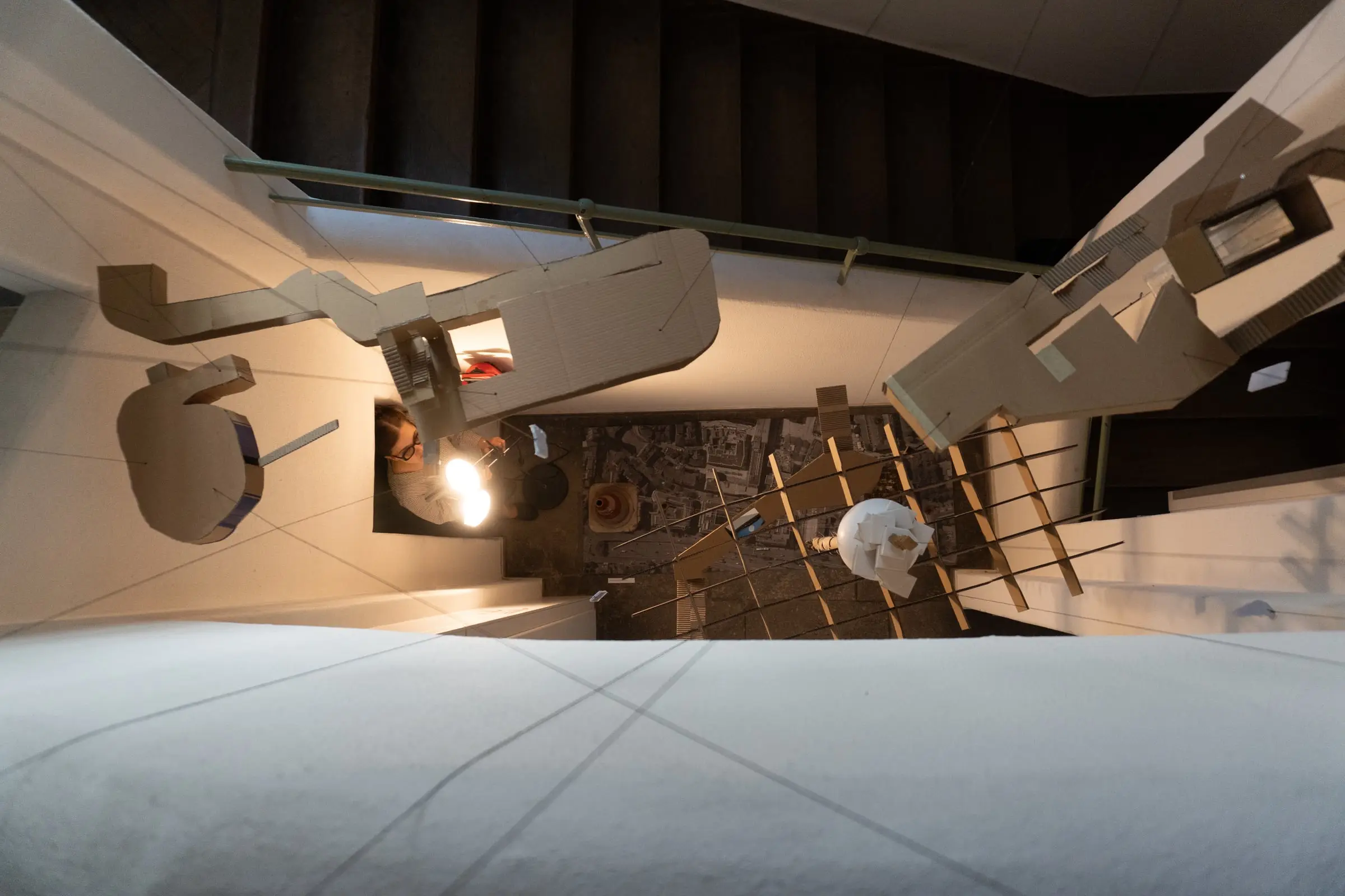 Looking straight down the stairwell onto the hanging model, a printed aerial map of the city on the floor below