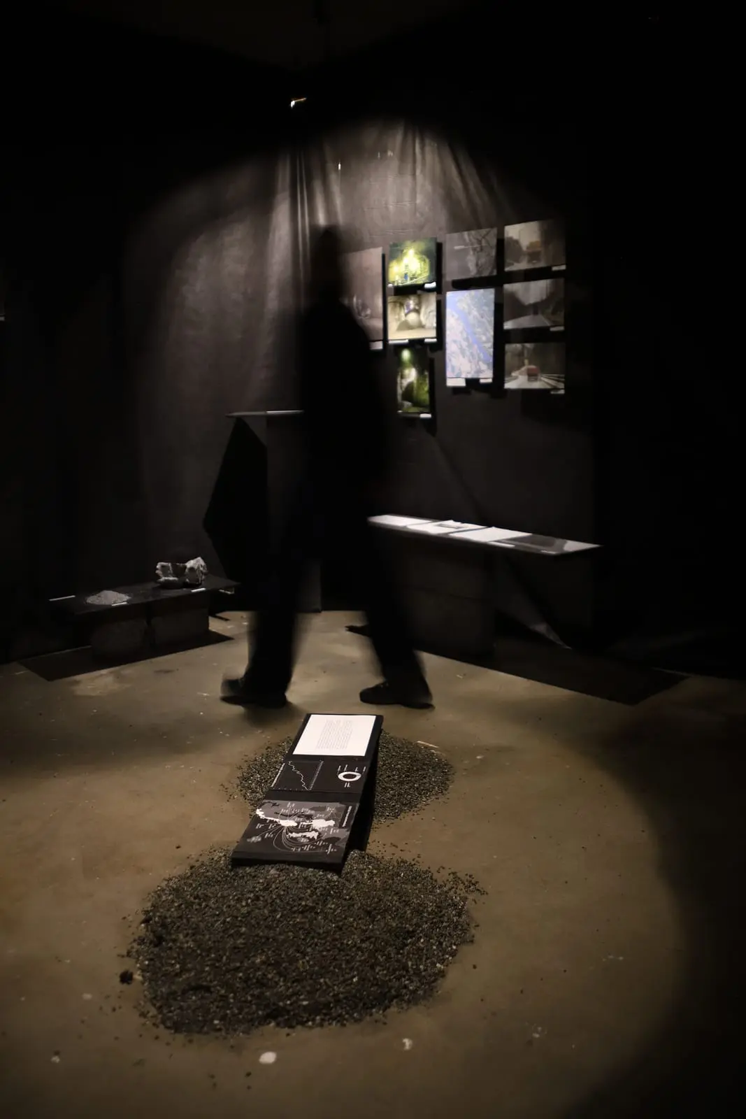 A blurred visitor walking past a heap of gravel with a folded booklet on it, photographs and benches against black fabric behind