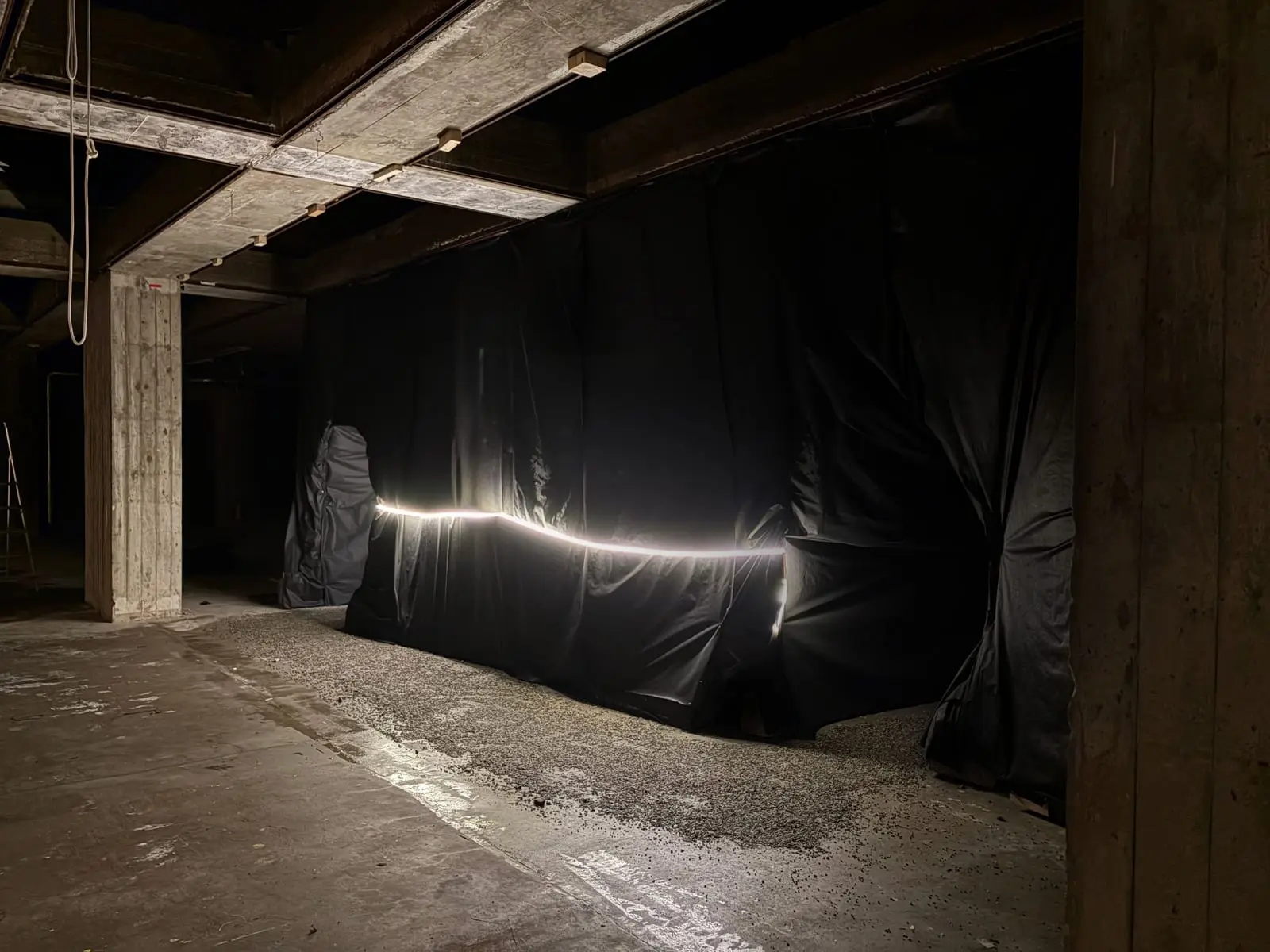 Black fabric hung between the concrete columns and beams of the existing hall, a line of light along it and gravel spread on the floor