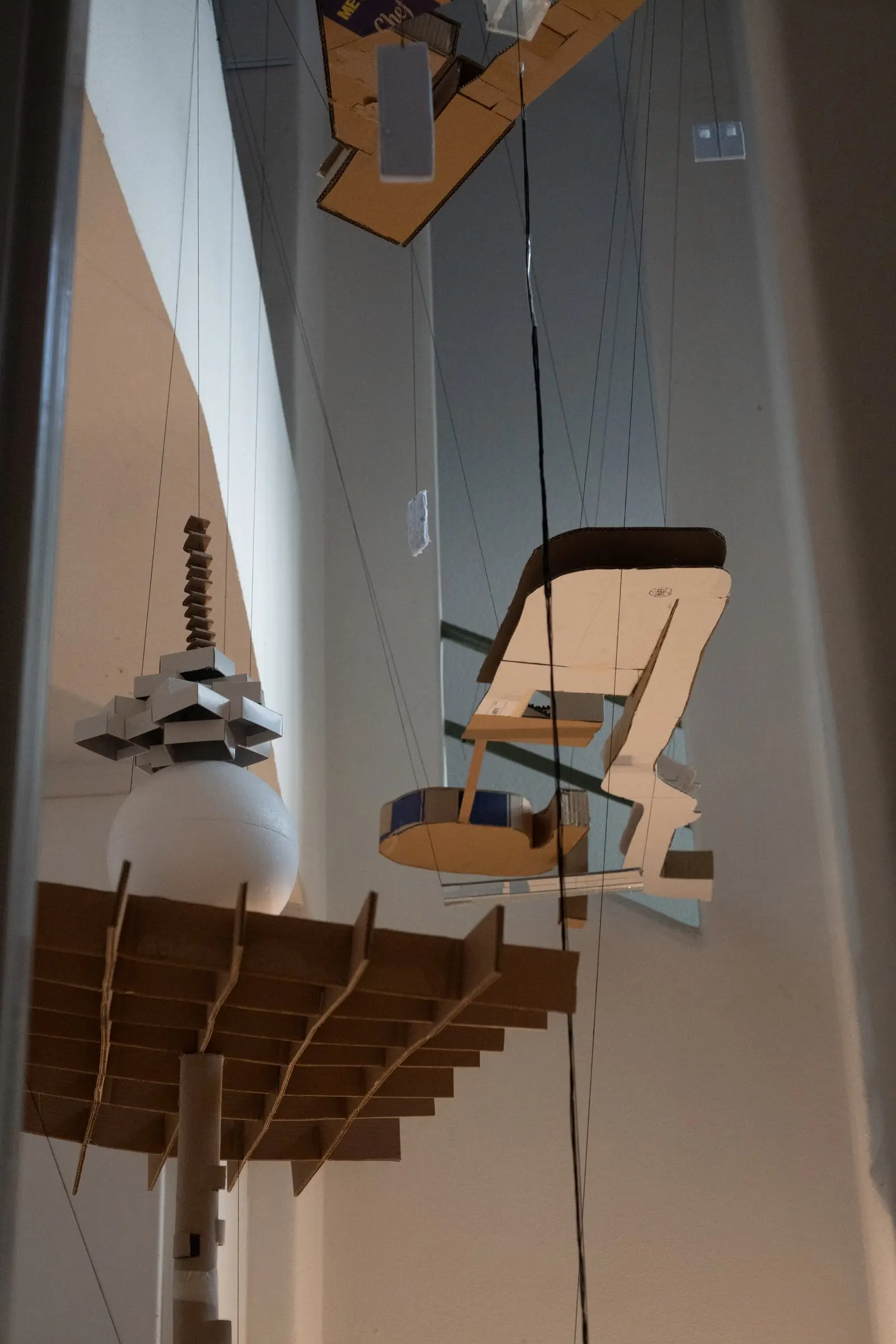 Several cardboard model pieces and a slatted grid hanging on wires in the stairwell