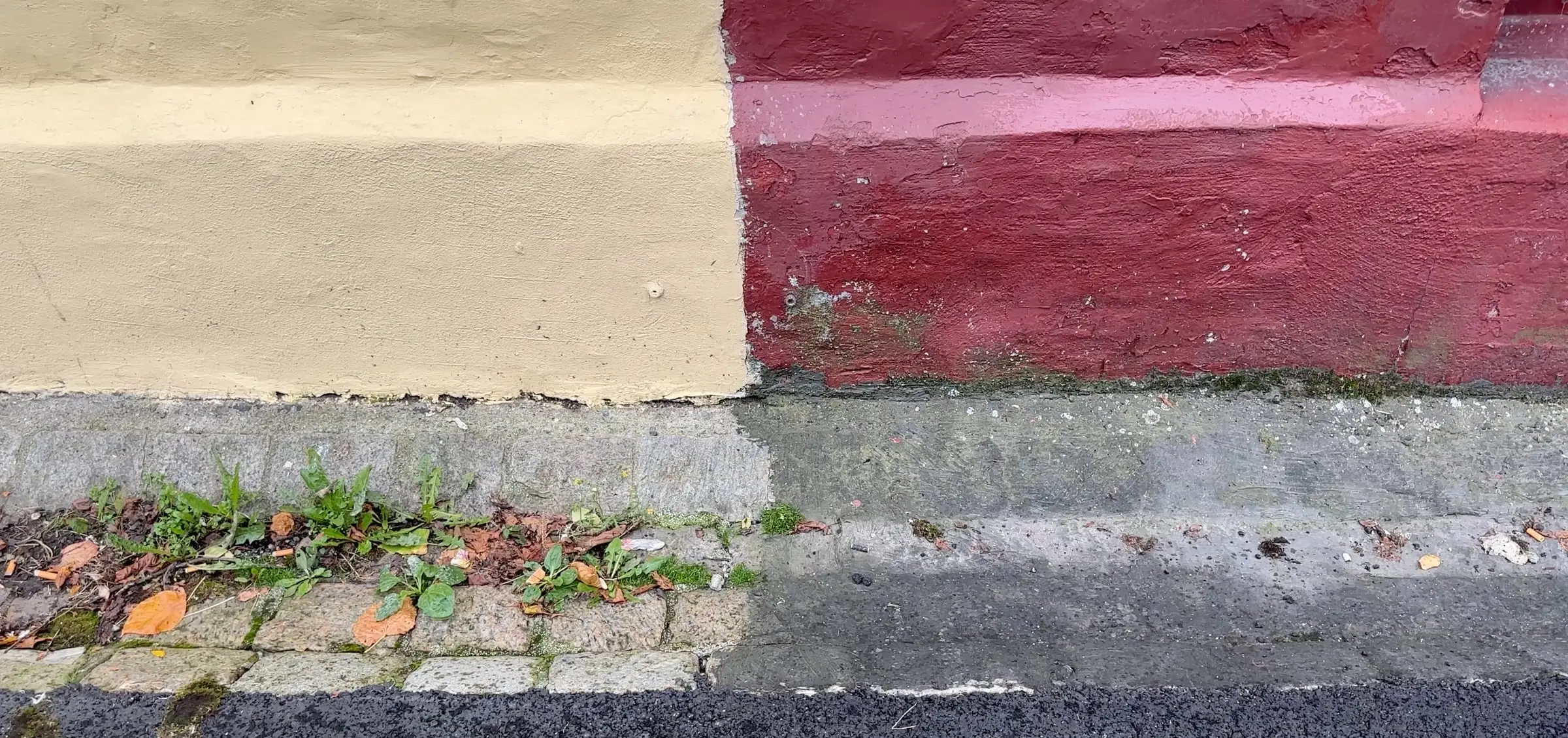 A pavement edge where asphalt, a concrete kerb and a strip of weeds meet the foot of a cream and red painted wall