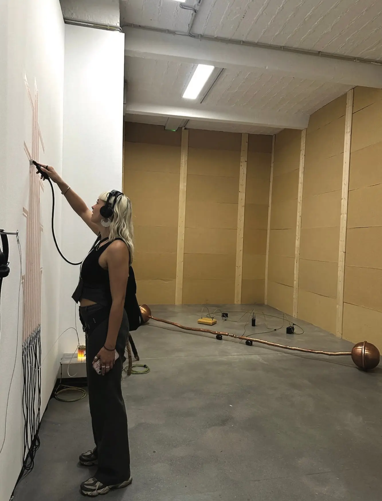 A visitor in headphones reaching up to a work on a white wall, insulated walls and a copper pipe on the floor behind