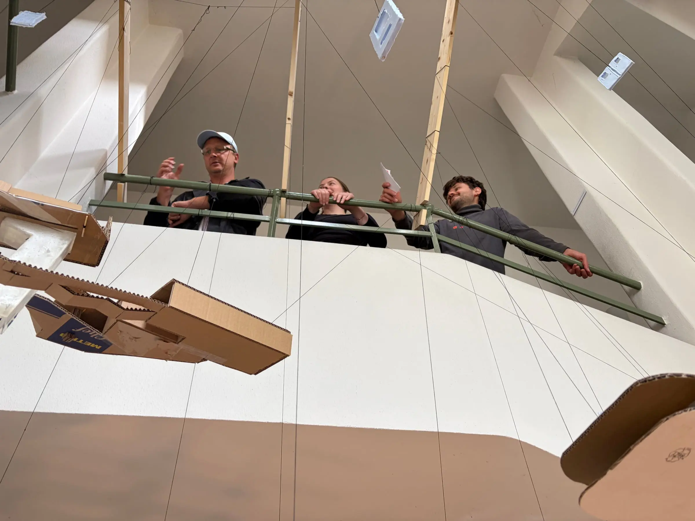 Three people at a balcony rail looking at the model hanging in front of them
