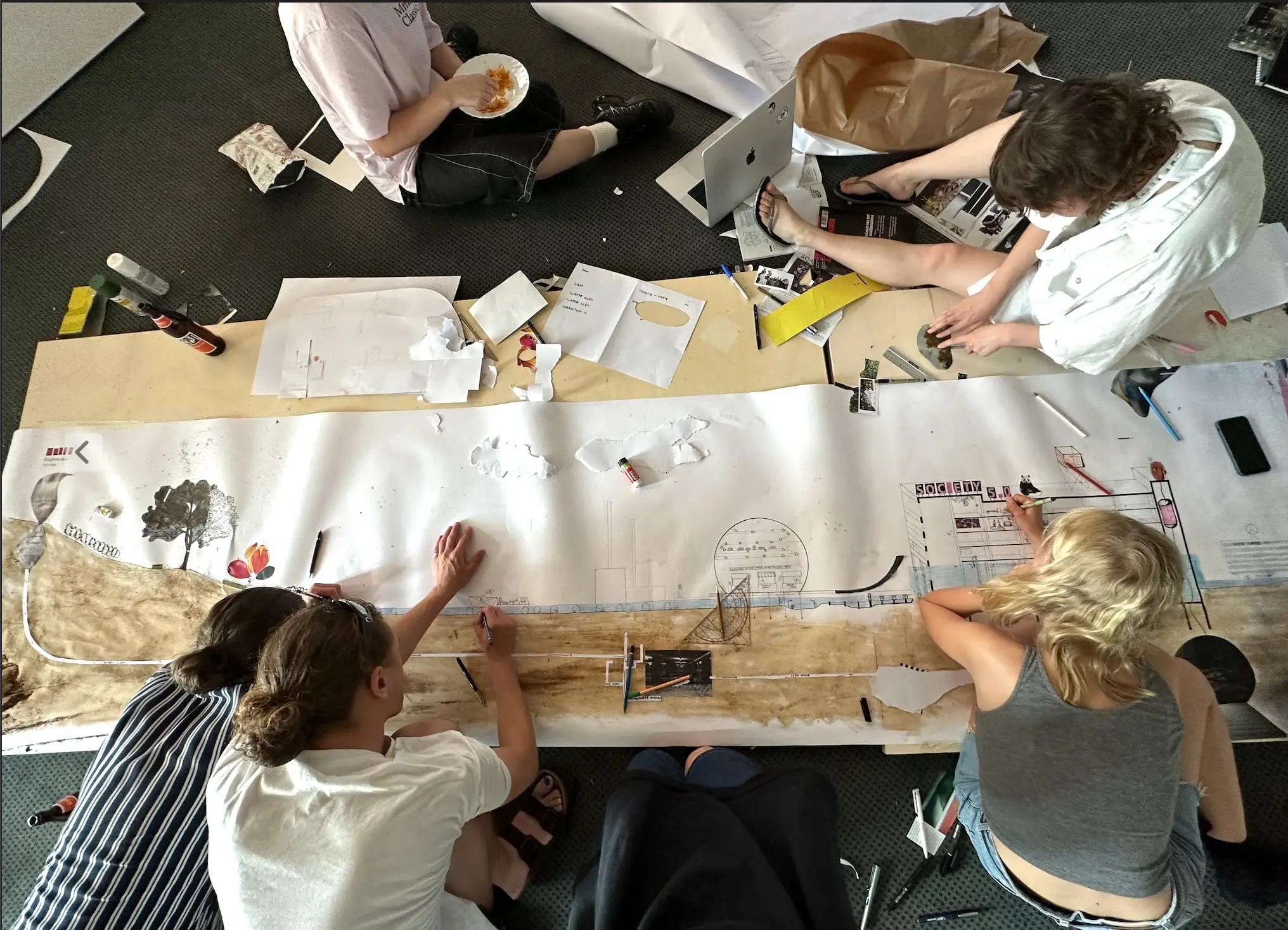Four students kneeling on the floor around the long paper section, drawing and cutting