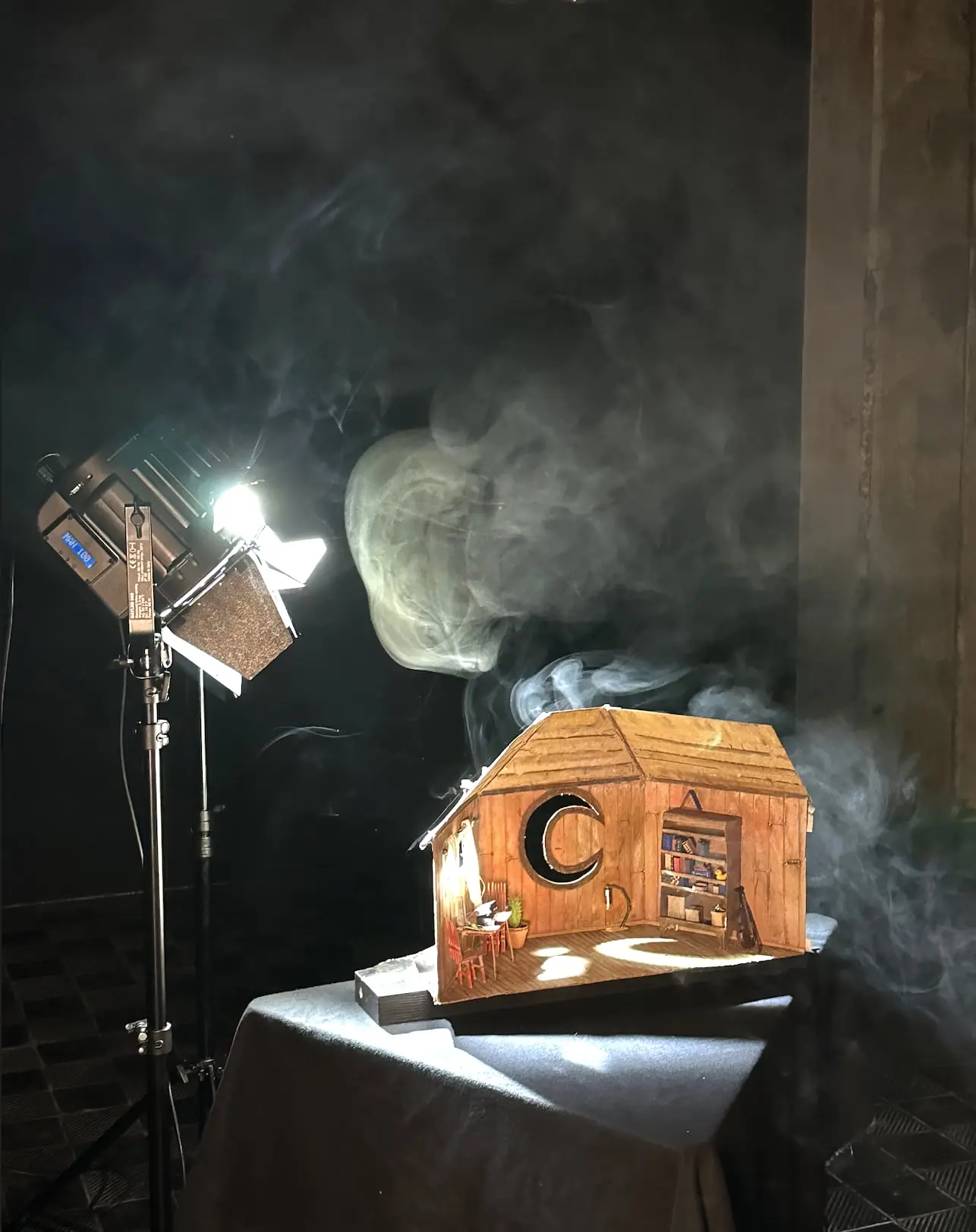 A small model house with a crescent moon on its wall, lit by a film lamp through smoke