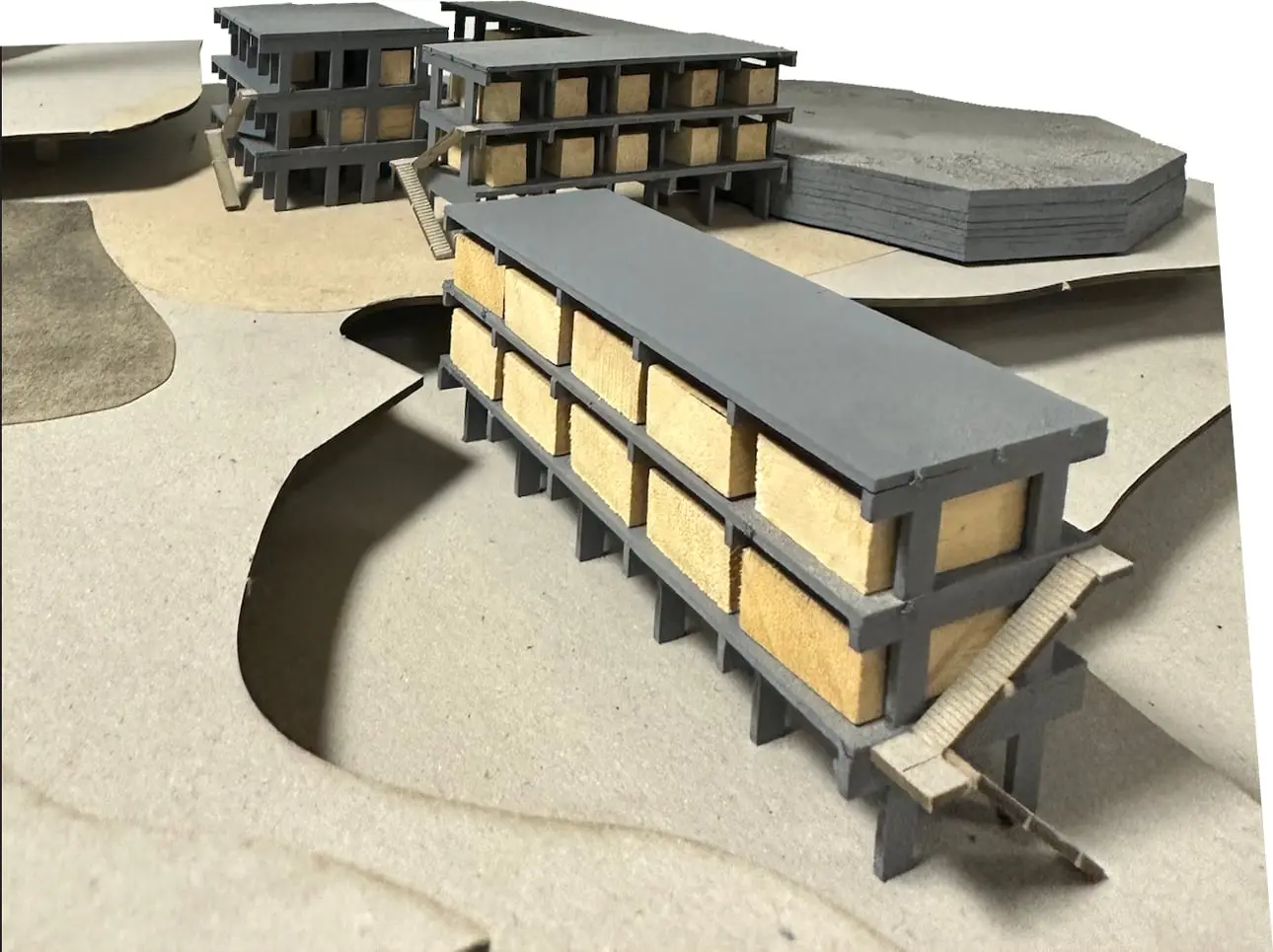 Model of a long grey concrete frame filled with pale timber boxes, standing over a cut landscape