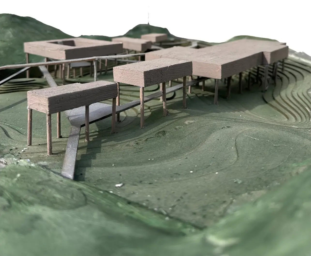 Model of timber-coloured buildings standing on thin columns over a green contoured landscape, joined by raised walkways