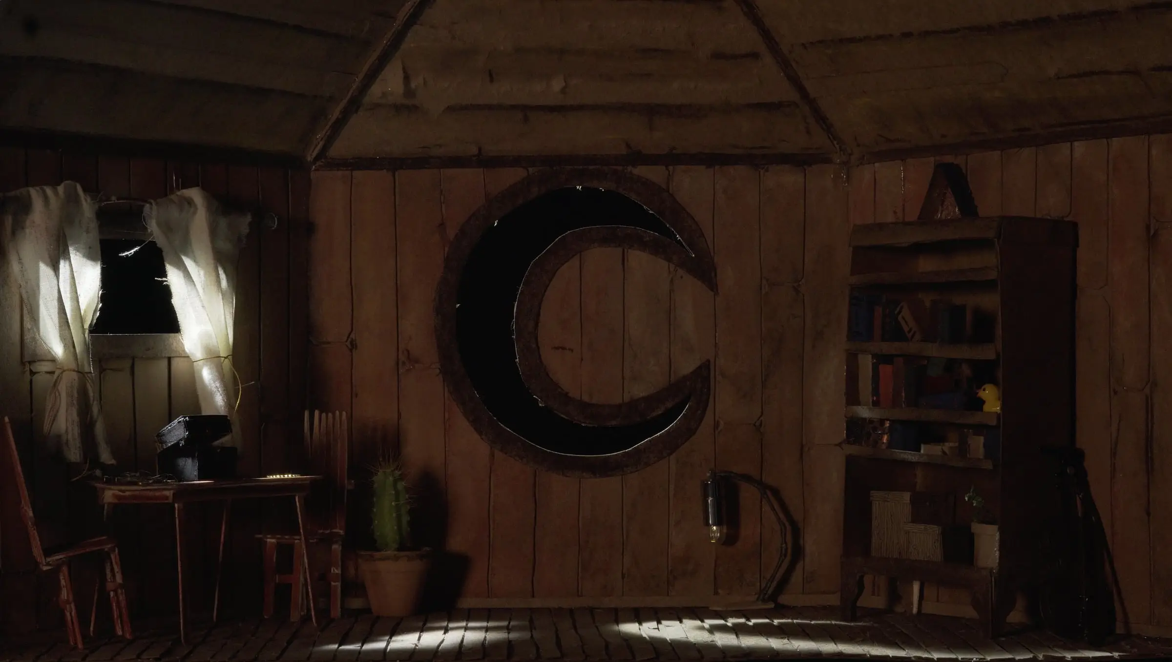 Dim wooden interior with a large crescent moon on the wall, a desk and a bookcase