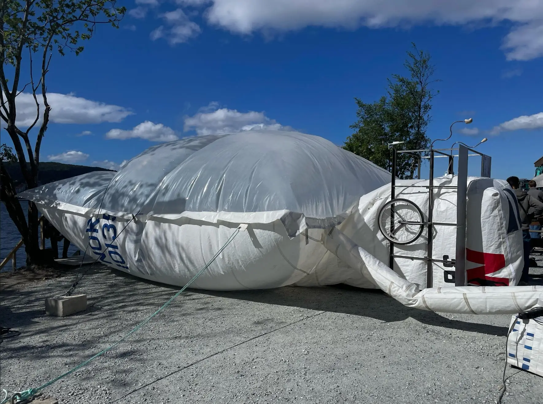 A long white inflatable of sail fabric and clear plastic lying on a quay under a blue sky, a fan unit at one end