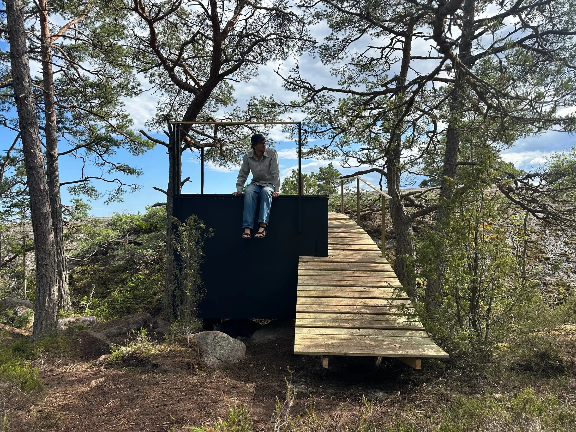 Someone sitting on the roof of the black shelter among pines, the curved timber bridge leading up to it