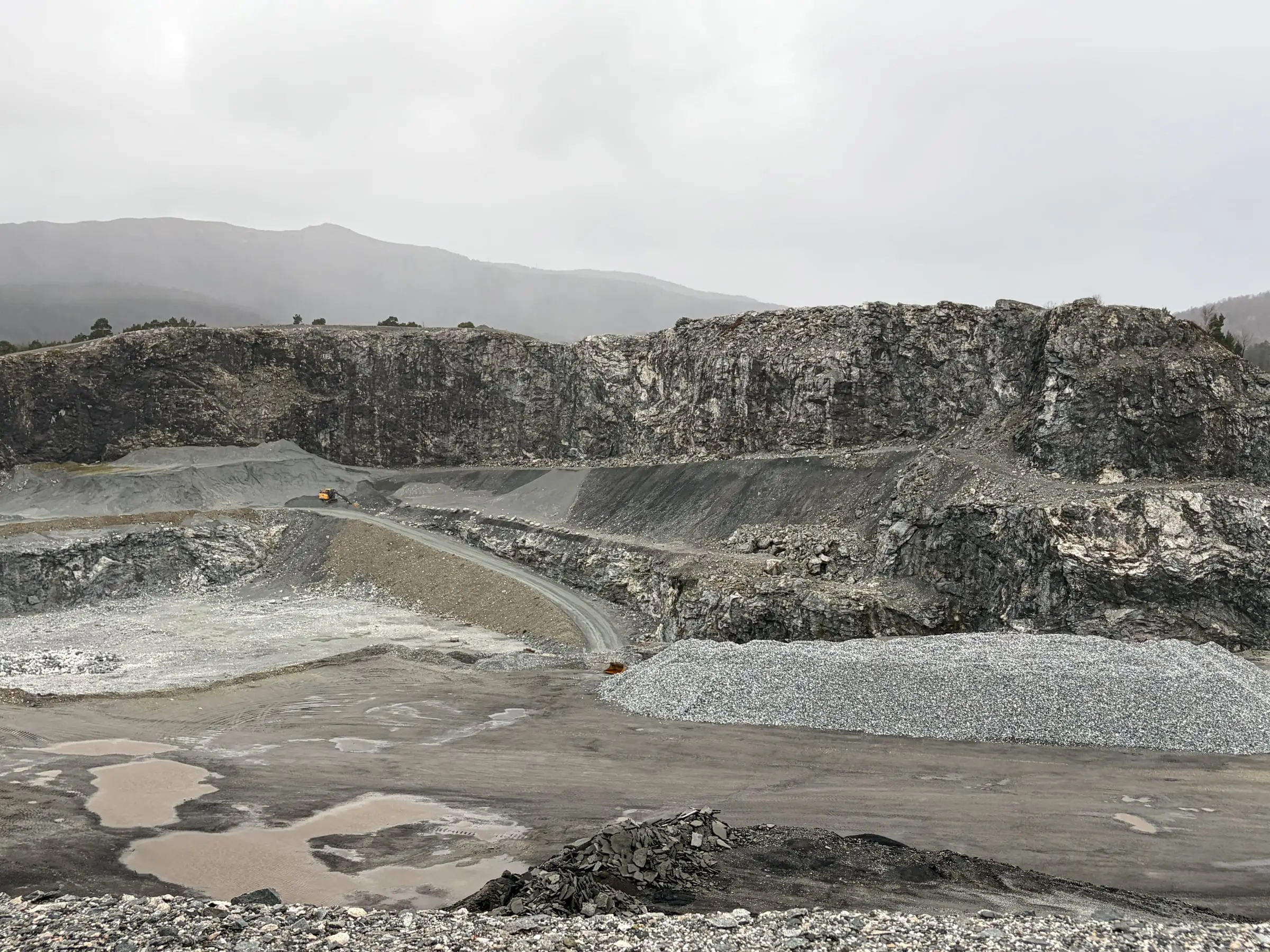 A wide quarry cut into a hillside, pools of water and heaps of gravel in front