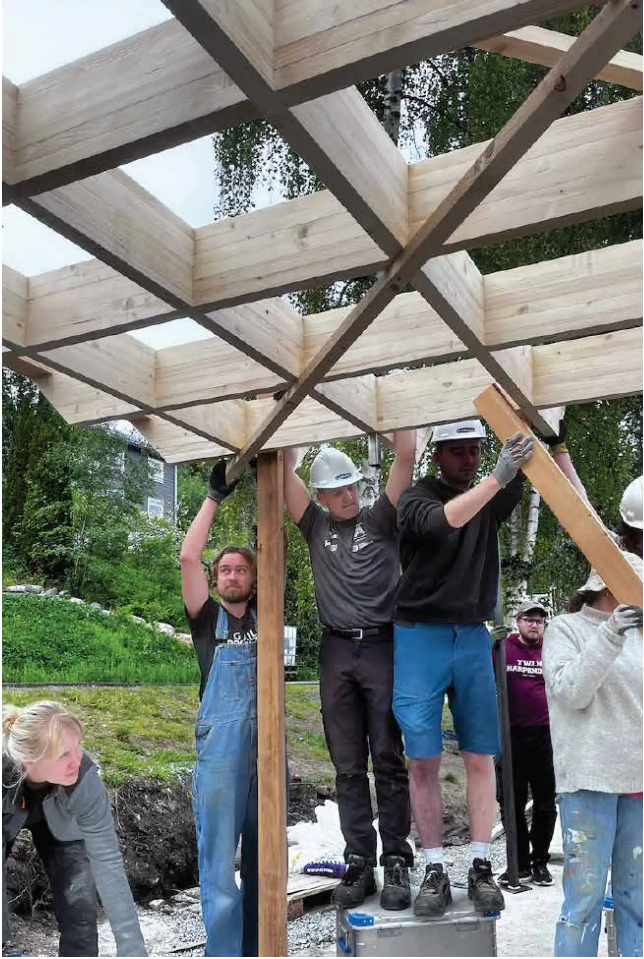 Students holding a timber post upright while the crossed roof beams are set above them