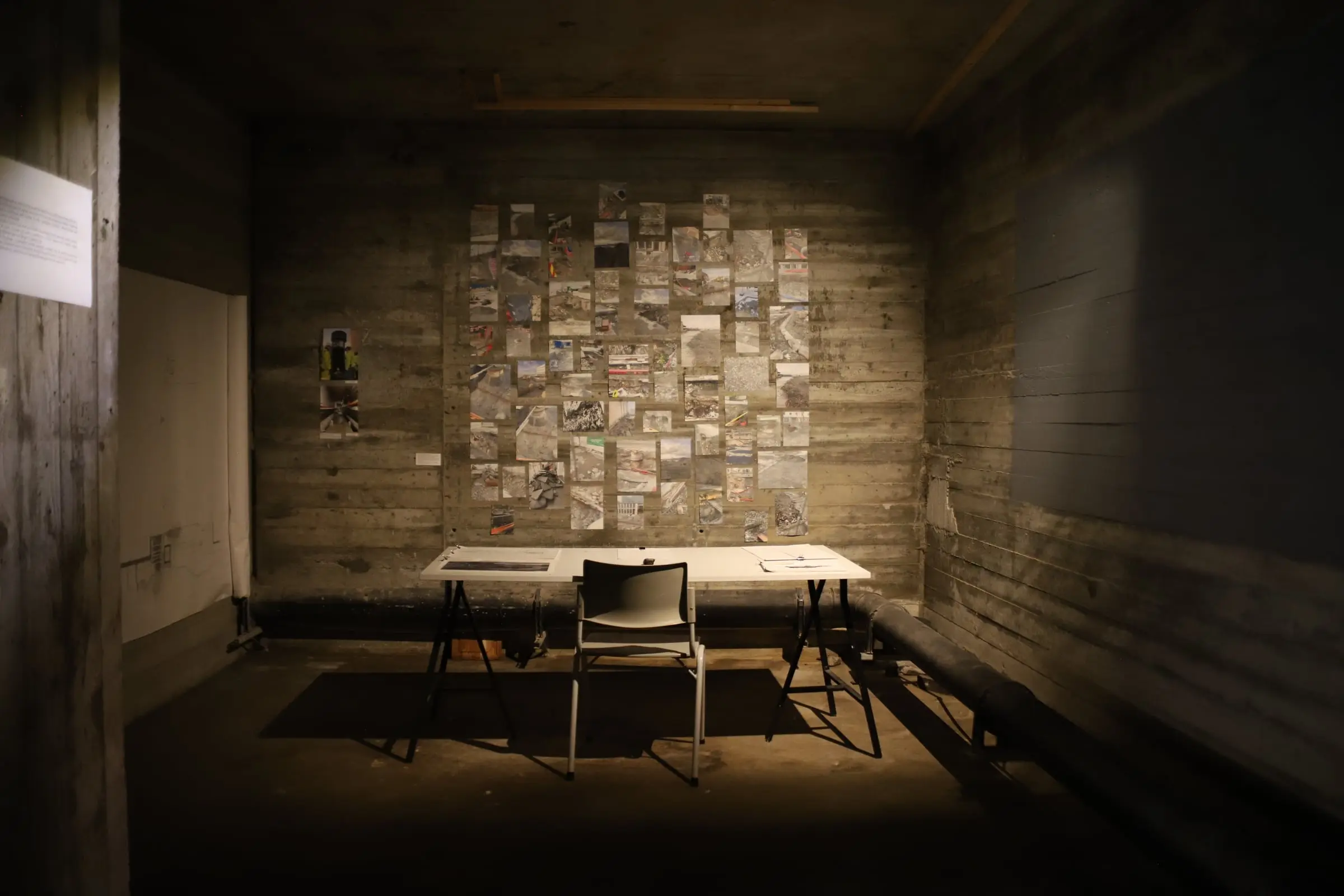 A wood-panelled room lit by a single lamp, a wall of pinned documents above a table and chair