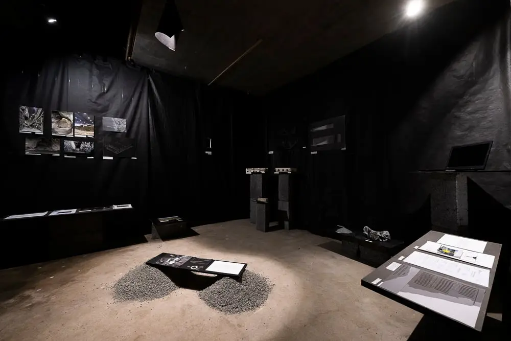 A dark room of black fabric with photographs on the walls, plinths, and a heap of gravel on the floor with a booklet on it