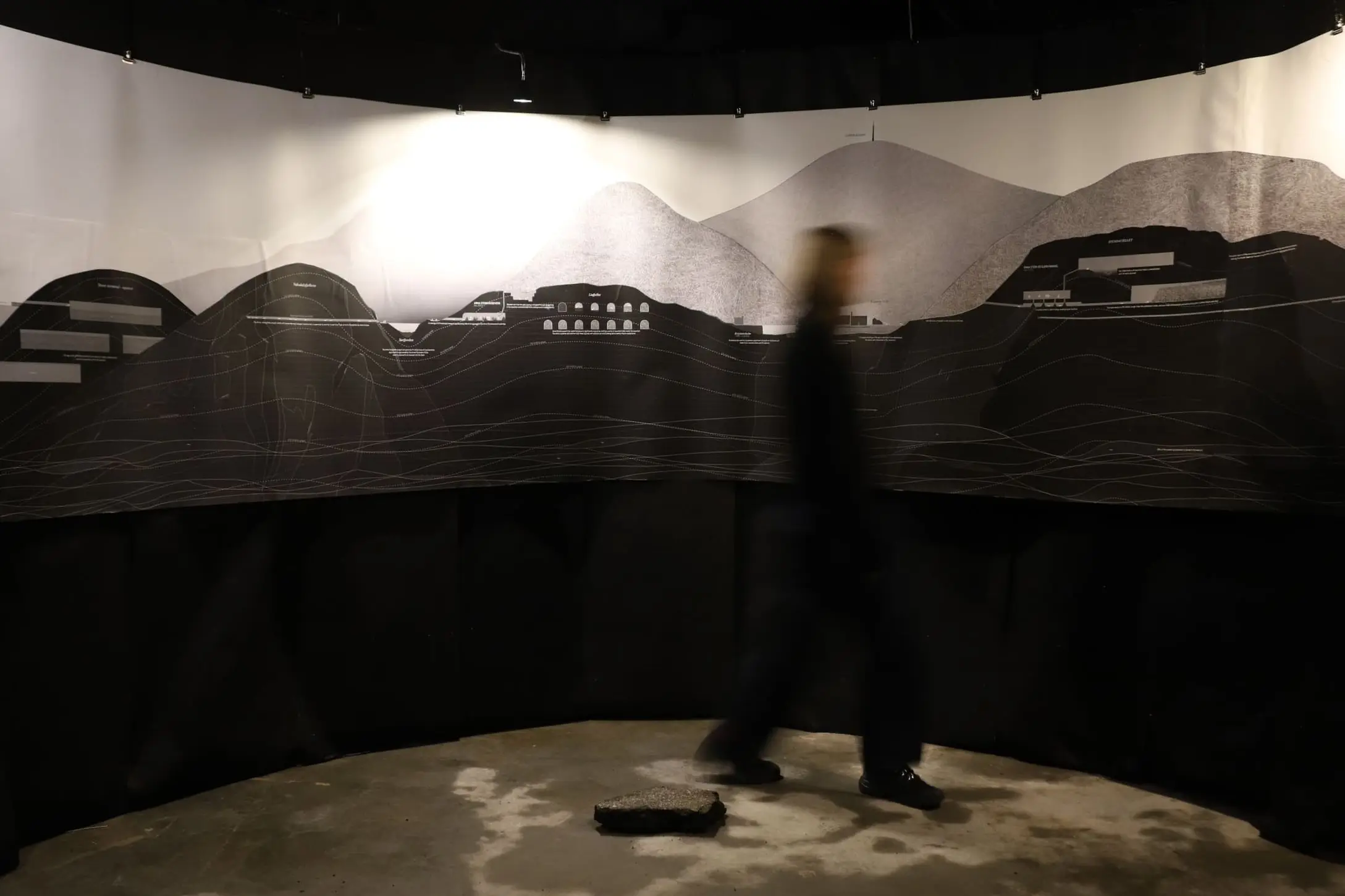 A blurred visitor crossing the round room, a printed mountain landscape curving around the wall above black fabric