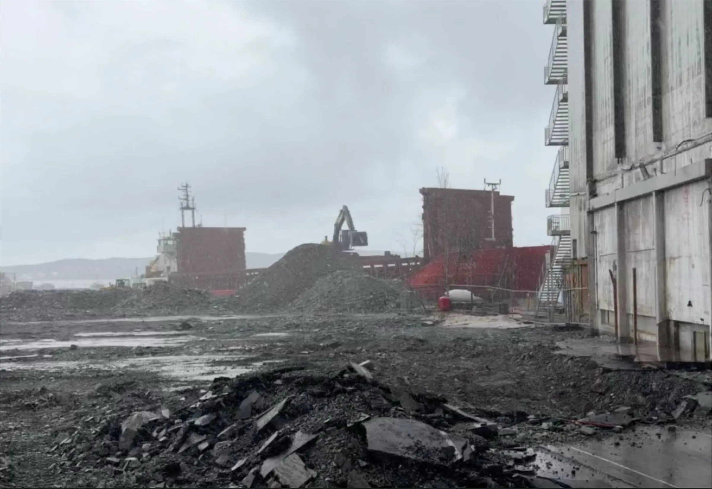 Heaps of rock and rubble on a wet harbour site, an excavator and ships behind