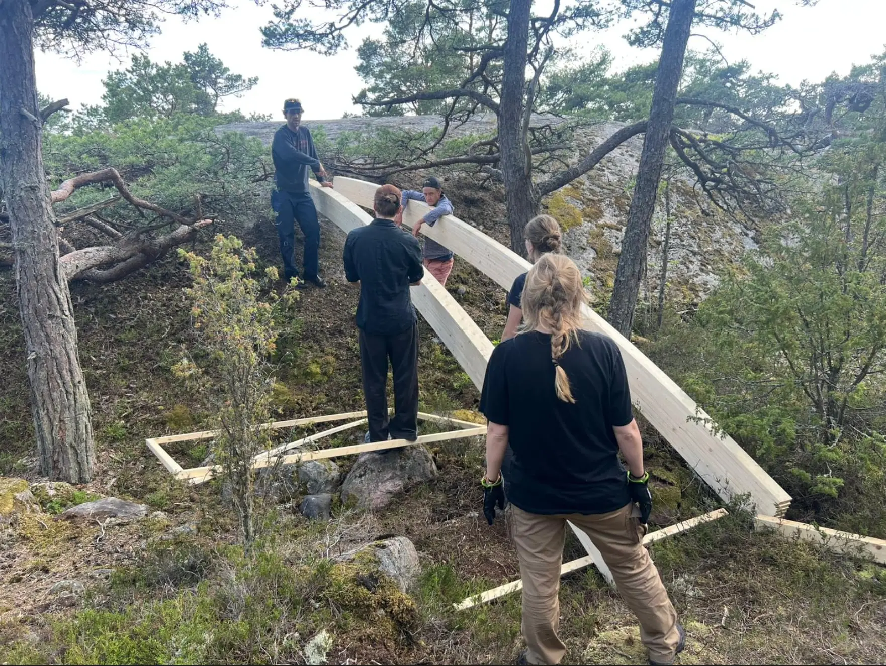 The team lifting a curved beam into place between pine trees on the rock