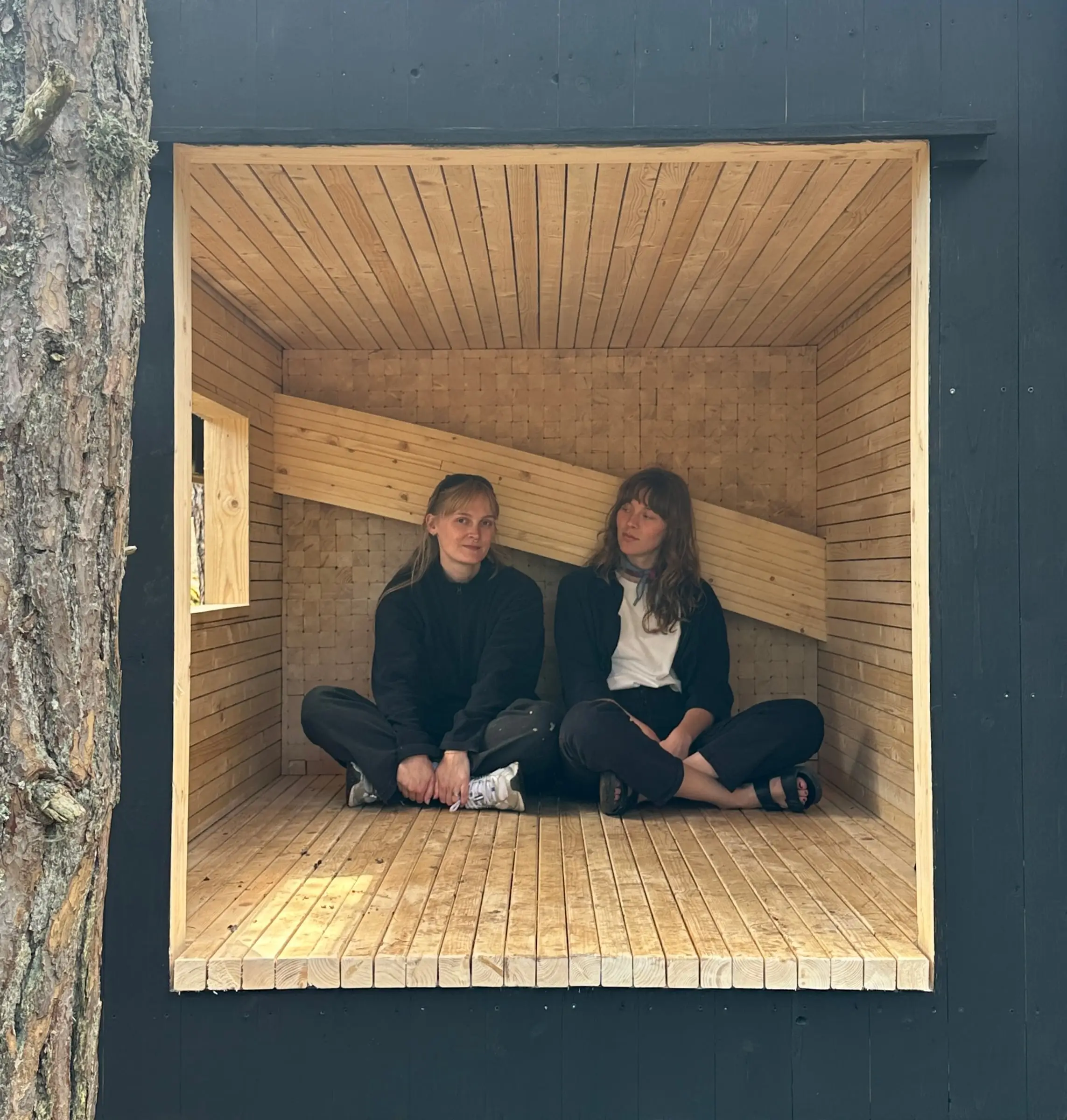 Two people sitting cross-legged inside the timber-lined shelter, seen through its square opening in the black facade