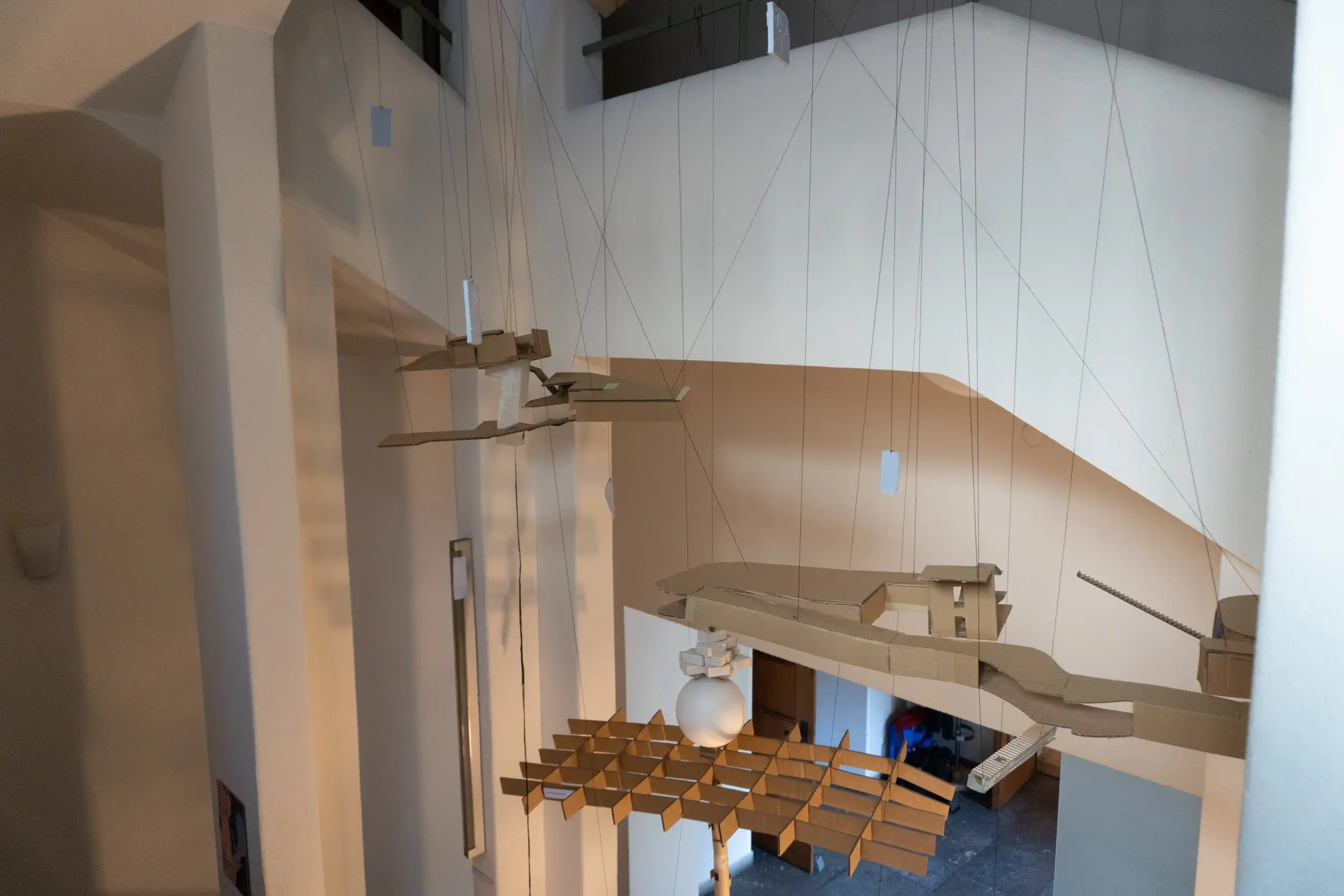A hanging model of cardboard, timber slats and a white sphere suspended through a white stairwell