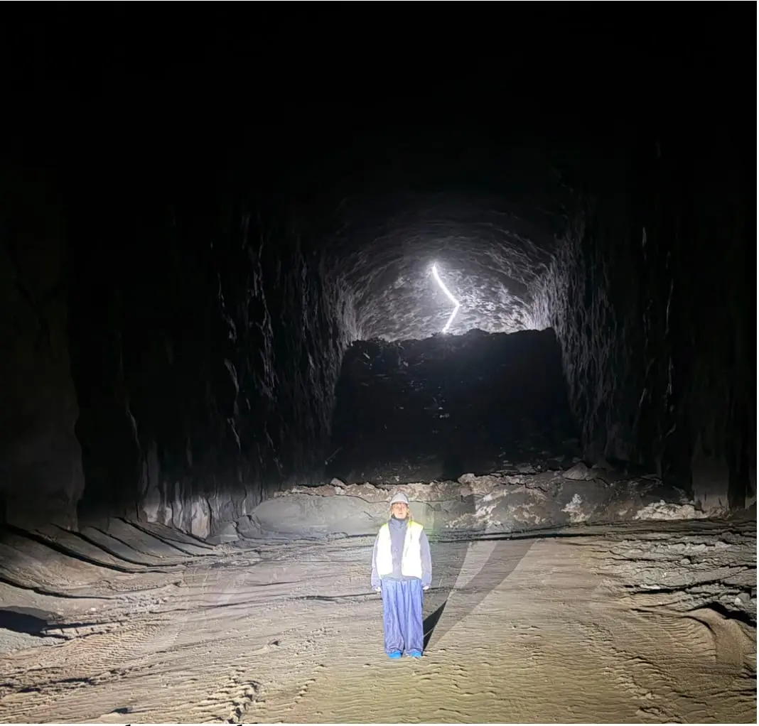 A small figure standing in a dark rock tunnel, light at the far end