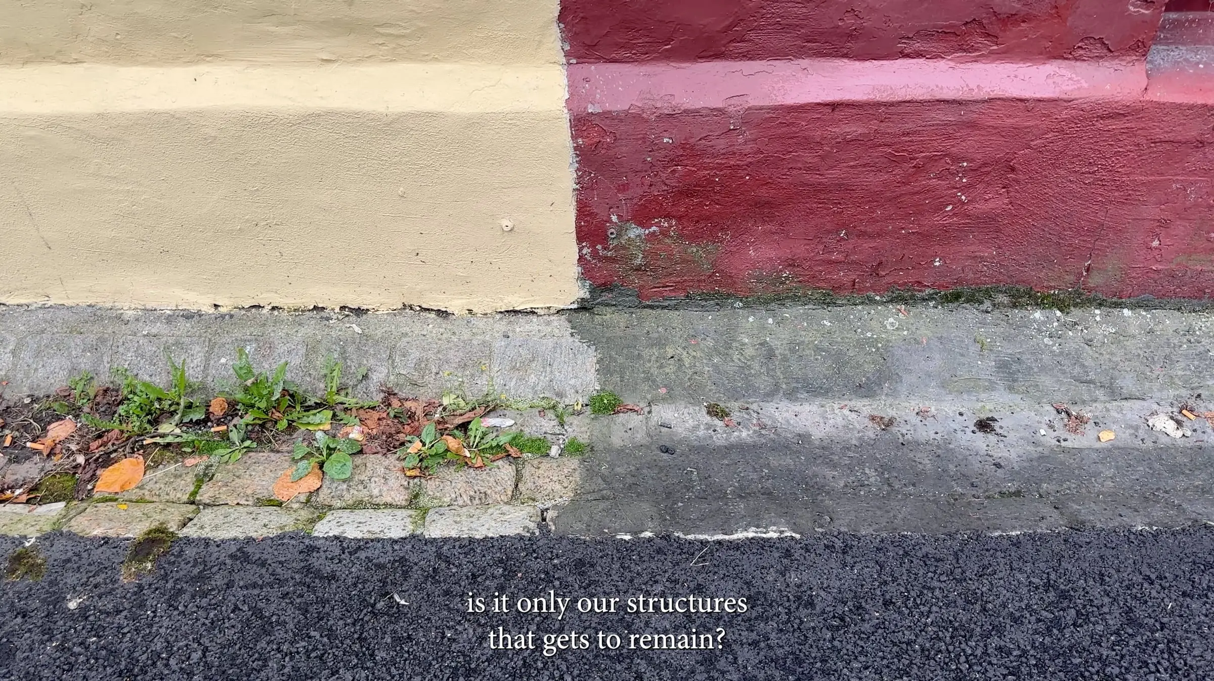 Film still of a pavement edge against a cream and red wall, with the subtitle 'is it only our structures that gets to remain?'
