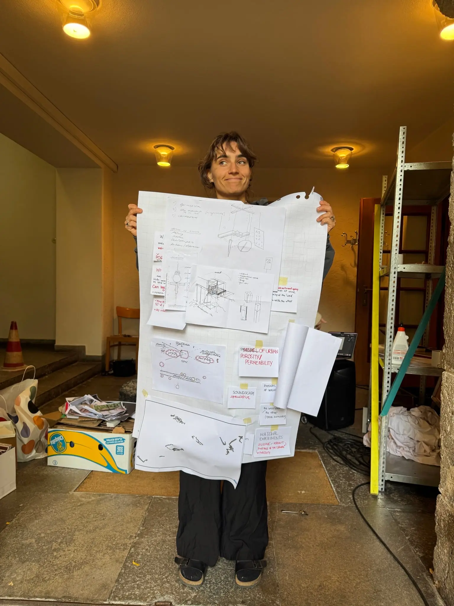 Someone holding up a large sheet covered in notes, sketches and photographs in a storage room